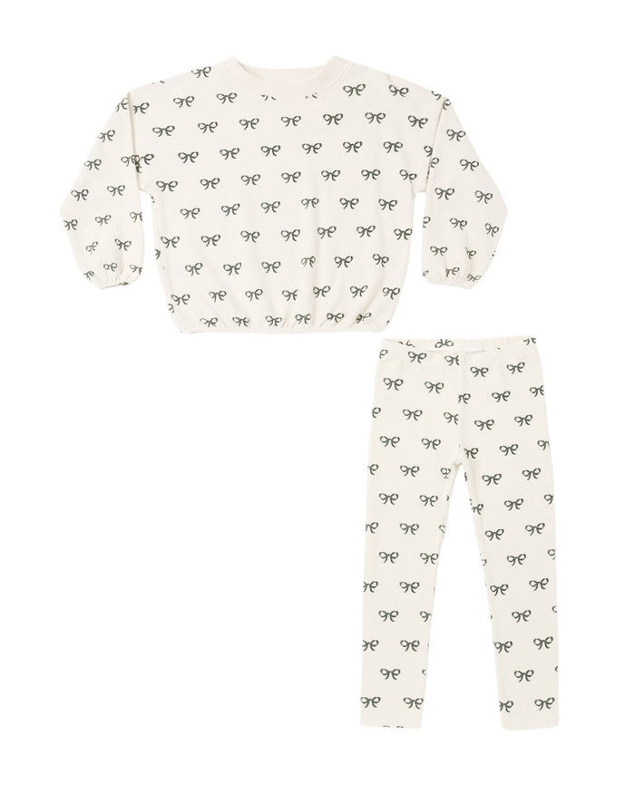 A two-piece children's pajama set featuring a long-sleeve top and matching leggings in an off-white color, both printed with a repeating pattern of small black bows.