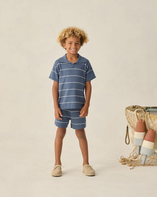 Child wearing a striped blue outfit standing next to a woven basket with beach balls on a beige background