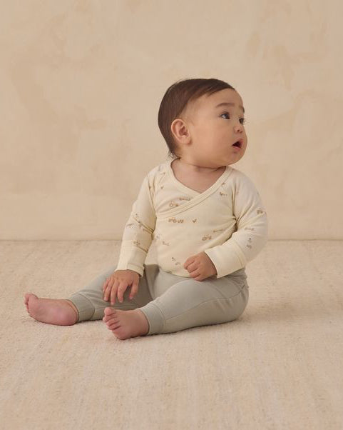 Baby sitting on a beige blanket wearing a light-colored long-sleeve shirt and gray pants.