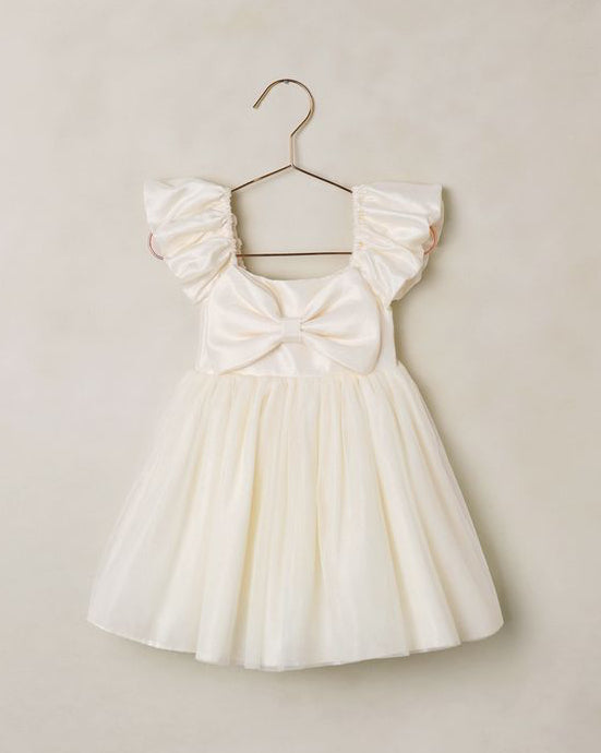 White dress with ruffled sleeves and a bow detail on a hanger against a beige wall.